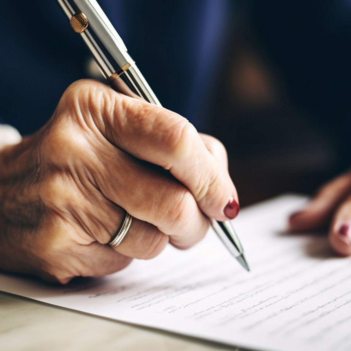 Die Hände einer älteren Dame sind zu sehen. In der Hand hält sie einen feinen Kugelschreiber und schreibt auf ein Blatt Papier.