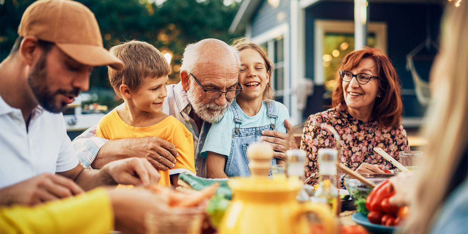 Ein glücklicher Großvater sitzt mit seiner Familie an einem Esstisch im Garten und hält seine Enkel im Arm