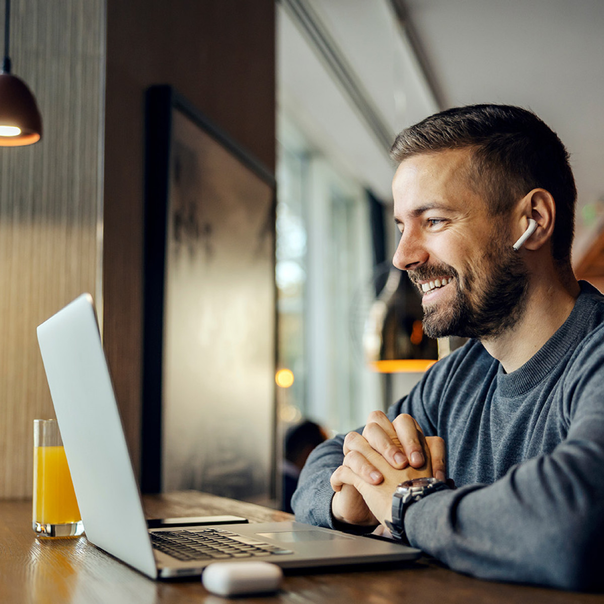 Ein Mann sitzt in einem Café am Tisch mit einem MacBook und einem Glas Orangensaft und führt eine Videokonferenz