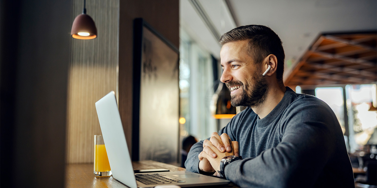 Ein Mann sitzt in einem Café am Tisch mit einem MacBook und einem Glas Orangensaft und führt eine Videokonferenz